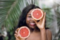 Amazing playful brunette african american woman with naked shoulders, holding halves of grapefruit in hands, hiding one