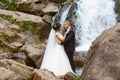 Amazing romantic view of happy bride and groom near beautiful grand waterfall in mountain. Success life. Marriage couple outdoor