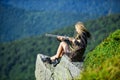 Amazon girl sit on cliff prepare gun for hunting. Aiming concept. Hunting season. Hunter mountains landscape background