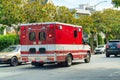 Ambulance truck as it speeds through downtown city streets in midday sun with trees and foliage in the background and other cars