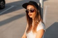 American cool girl in black fashion cap in vintage sunglasses in summer clothes resting on street at sunset. Portrait beautiful Royalty Free Stock Photo