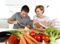 American couple working in domestic kitchen together wife following recipe in cookbook and husband slicing cheese