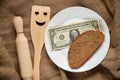 American dollar and a piece of black bread on a white plate and next to it lies a kitchen wooden spatula with a smiley face and a