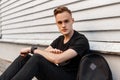 American fashionable young man in a summer black stylish clothes with a backpack sitting in the city on the sidewalk near a wall Royalty Free Stock Photo