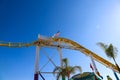 An American flag and a California flag on top of a yellow rollercoaster with a gorgeous blue sky and tall lush green palm trees