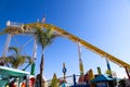 An American flag and a California flag on top of a yellow rollercoaster with a gorgeous blue sky and tall lush green palm trees