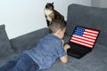 American flag on a laptop display, a little schoolboy in jeans lies on a sofa and scrolls, a cat sits nearby, a concept of