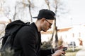 American good-looking young man hipster in sunglasses in black clothes in fashion baseball cap with backpack with mobile phone Royalty Free Stock Photo