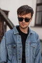 American handsome young man with a beard in fashionable sunglasses in a stylish blue denim jacket in a t-shirt stands Royalty Free Stock Photo