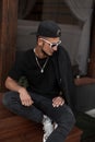 American hipster young man with a beard in a cap in a T-shirt in a jacket in sunglasses sits on wooden stairs near a vintage cafe Royalty Free Stock Photo