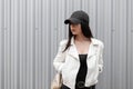 American pretty young brunette woman in a trendy white leather jacket in a stylish black baseball cap in a T-shirt Royalty Free Stock Photo