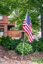 American stars and stripes flag on pole stuck in ground in front of rustic landscaped home under spreading green tree branches