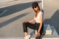 American stylish girl hipster in fashion black cap in sexy white top in trendy jeans and sneakers rest on asphalt outdoors in Royalty Free Stock Photo