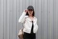 American stylish young woman in a trendy black baseball cap in a leather white jacket in black jeans with a gold backpack Royalty Free Stock Photo
