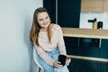 American teenage girl at Home kitchen interior holding Black coffee cup. Woman in pink blouse and blue jeans sitting on white chai Royalty Free Stock Photo