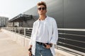 American young handsome man fashion model in white stylish shirt in blue vintage jeans in trendy sunglasses is posing Royalty Free Stock Photo