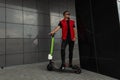 American young hipster man with a hairstyle in stylish denim red-black clothes in sunglasses stands with a modern electric scooter Royalty Free Stock Photo