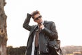 American young hipster man in stylish dark sunglasses in a trendy leather black jacket with a backpack posing near a wall in the Royalty Free Stock Photo