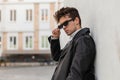 American young hipster man in a trendy leather black jacket rests near a wall in the city on spring day. Cool guy straightens Royalty Free Stock Photo