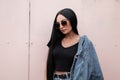 American young hipster woman in stylish sunglasses in a black top in a fashionable blue denim jacket in a trendy skirt stands Royalty Free Stock Photo