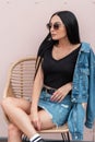 American young hipster woman in sunglasses in a black top in a blue denim jacket in a skirt is sitting on a straw chair Royalty Free Stock Photo