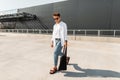 American young man fashion model in trendy jeans in a stylish shirt in fashionable sandals in sunglasses with bag walks near a Royalty Free Stock Photo