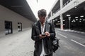 American young man in leather black jacket with backpack with hairstyle with modern mobile phone stands in the city. Handsome