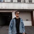 American young man in stylish sunglasses in a vintage blue denim jacket in a black T-shirt stands near a building on the street. Royalty Free Stock Photo