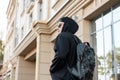 American young man tourist in a stylish black hooded sweatshirt with a hood with trendy leather backpack travels through the city Royalty Free Stock Photo