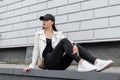 American young woman in a jacket in leather sneakers in black jeans in a stylish baseball cap with a backpack sits on a tile Royalty Free Stock Photo