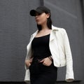 American young woman in a white fashionable leather jacket in black jeans in a stylish black cap in a T-shirt is standing Royalty Free Stock Photo