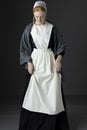 An Amish woman wearing a long black dress, shawl, apron, and cap against a studio backdrop