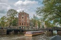 Tree-lined canal with bridge, touristic boat, buildings and cloudy sky in Amsterdam.