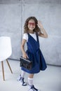 Little girl in red eyeglasses, wearing in dress, holding a bag in hand, looking at camera, over concrete background