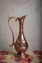 An ancient copper jug with hand-made patterns and ornaments standing on a table with a carpet with national patterns and prints.