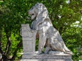 Ancient marble white lion statue with shield and heraldic symbols. Against background of green trees with abundant foliage.