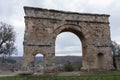 An ancient, weathered stone arch with two openings stands against a backdrop of a cloudy sky and distant landscape