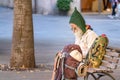 A senior man backpacker sitting on a square bench, is writing down notes on a notepad wearing eyeglasses and a gnome wool hat