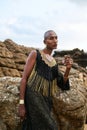 Androgynous ethnic fashion model in luxury dress sits on rocks by ocean. Gay black person in jewellery and posh clothes