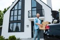 Angle view of cheerful man holding boxes near car and modern house Royalty Free Stock Photo