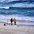 Angler in warm winter clothes with attached hoods and hands in pockets hold the fishing rod in the Mediterranean Sea on the beach