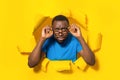 Angry african american man looking at camera through eyeglasses, breaking through torn hole in yellow paper Royalty Free Stock Photo