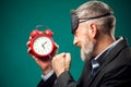 Angry bearded businessman with sleep mask on head holding alarm clock, shouting and showing fist at it. Time management concept Royalty Free Stock Photo