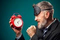 Angry bearded businessman with sleep mask on head holding alarm clock, shouting and showing fist at it. Time management concept Royalty Free Stock Photo