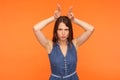 Angry bully brunette woman in denim dress showing bull horns with fingers on head, antler gesture