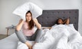 Angry Caucasian woman sitting on bed, covering her ears with pillow, suffering from her husband& x27;s snoring at home Royalty Free Stock Photo