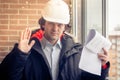 An angry disgruntled builder worker in a helmet with project drawings plans in his one hand and mobile phone in another Royalty Free Stock Photo