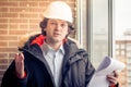 An angry disgruntled builder worker in a helmet with project drawings plans in his one hand and mobile phone in another