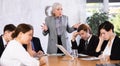 Angry elderly female boss scolding office workers of different ages using notebooks at table in office