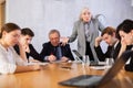 Outraged old female manager getting mad at office workers sitting with reports and laptops at table in meeting room